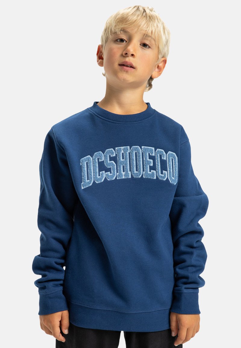 Námornícky modrý sveter vyrobený z mäkkej tkaniny, s výrazným logom "DCSHOECO" v svetlomodrej farbe cez hruď. Dlhé rukávy, dizajn s okrúhlym výstrihom.