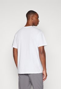 Tricou alb din bumbac, cu guler rotund, mâneci scurte și o croială lejeră, văzut din spate pe un fundal deschis.