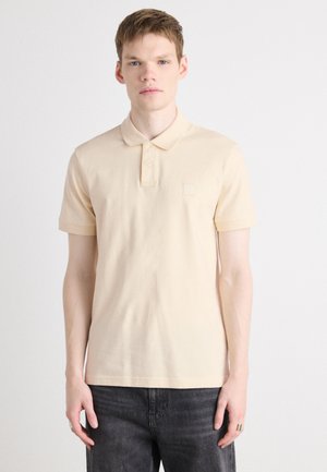 Lys beige polo-skjorte med klassisk krage, korte ermer og et diskret logo-patch på brystet. Mykt bomullsmateriale med en jevn tekstur.