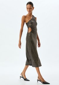Asymmetrisches Midi-Kleid aus schwarzem und goldenem Paillettenstoff mit Raffung und seitlichen Ausschnitten, kombiniert mit schwarzen Slingback-High Heels. Glatte Textur, figurbetonte Form.