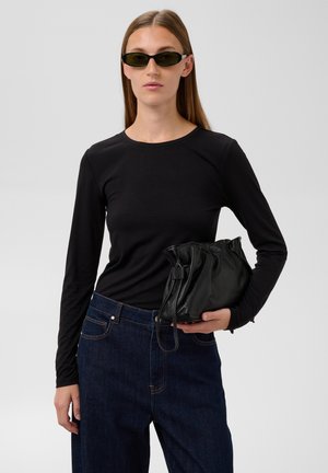 Une femme porte des lunettes de soleil noires, un haut noir à manches longues et un jean foncé. Elle tient un sac souple noir à cordon devant un fond gris uni.