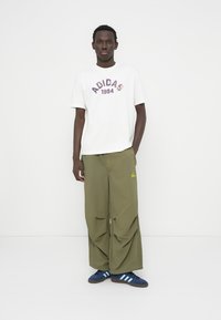 Λευκό μπλουζάκι με τη φράση «ADIDAS 1994» σε μοβ, φαρδιές παντελόνες ελιάς με τσέπες μπροστά και μπλε παπούτσια με τρεις λευκές ρίγες.