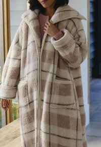 Kostium w kratę z polaru w kolorze kremowym i delikatnym różu, z suwakiem, dużymi kieszeniami i szerokim kołnierzem. Przyjemna w dotyku tekstura i luźny krój.