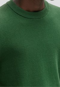Pull en tricot vert avec un col rond. Le matériau a un motif texturé et une finition côtelée autour du col. La vue rapprochée met en valeur le détail du tissu.