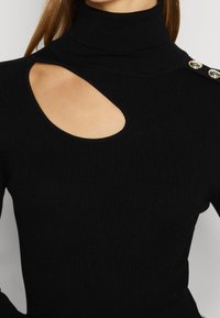 Pull à col roulé côtelé noir avec une découpe en forme de goutte sur le haut de la poitrine et deux boutons métalliques sur l'épaule gauche.