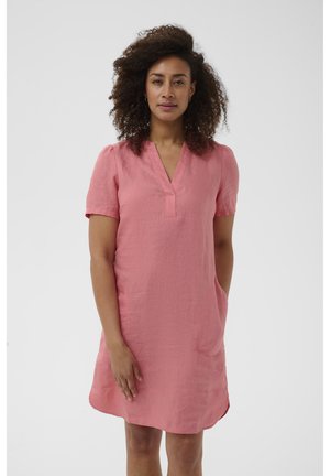 Femme aux cheveux bouclés portant une robe rose à manches courtes, longueur genou, debout avec une main dans la poche devant un fond uni.