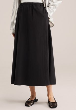 Modelka nosi dlhú čiernu sukňu s elastickým pásom, biely top, pruhovanú košeľu a čierne tkané baleríny, stojaca pred béžovým pozadím.