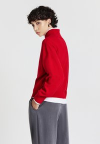 Maglione rosso a collo alto realizzato in morbido tessuto knit, con una vestibilità comoda e orlo a costine. Indossato sopra una camicia bianca con colletto; pantaloni grigi.