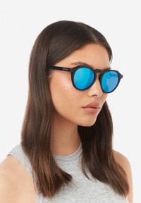 Sluneční brýle s černým rámem a modrými zrcadlovými čočkami. Mají jedinečný geometrický tvar a tenké nožičky. Hladká matná textura.