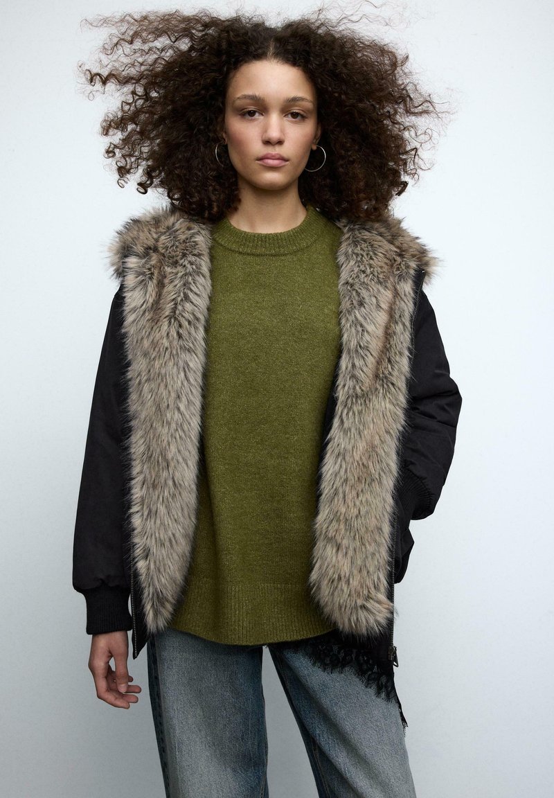 Czarna kurtka bomberka z kołnierzem z futerka syntetycznego, pod spodem oliwkowozielony sweter dziany, oraz jasnobrązowe dżinsy z koronkową lamówką na dole.