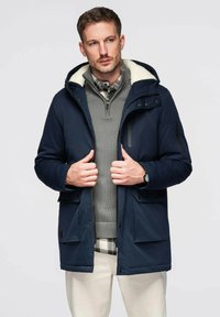 Veste bleu marine avec une capuche doublée de fausse fourrure, fermeture éclair à l'avant et poches latérales ; portée sur un pull gris et une chemise à carreaux.