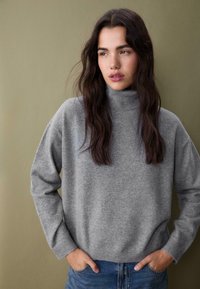 Siv turtleneck pulover iz mehkega pletenega materiala z sproščenim krojem. Dolgi rokavi in ribast rob dopolnjujeta sproščen dizajn.