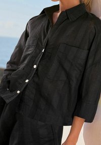 Kvinde iført en sort struktureret skjorte med knaplukning og trekvartærmer samt matchende bukser, der står op ad en lys væg med havet i baggrunden.