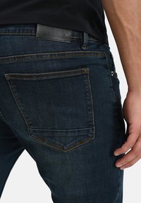 Los pantalones vaqueros de mezclilla azul oscuro presentan un clásico diseño de cinco bolsillos, costuras naranjas en contraste y un parche de cuero negro en la parte trasera de la cintura.