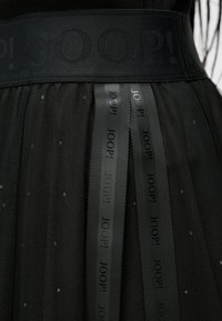 Melna tilla svārki ar rakstainu jostu, uz kuras izcelts uzspiests "JOOP!" logo un kontrastējošām atlas auduma lentēm. Audumā nelieli mirdzumi.