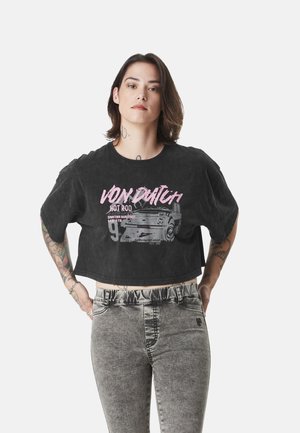 Personne aux cheveux mi-longs et foncés portant un T-shirt court noir avec le texte rose « Von Dutch » et un jean taille haute gris, debout les mains sur les hanches.