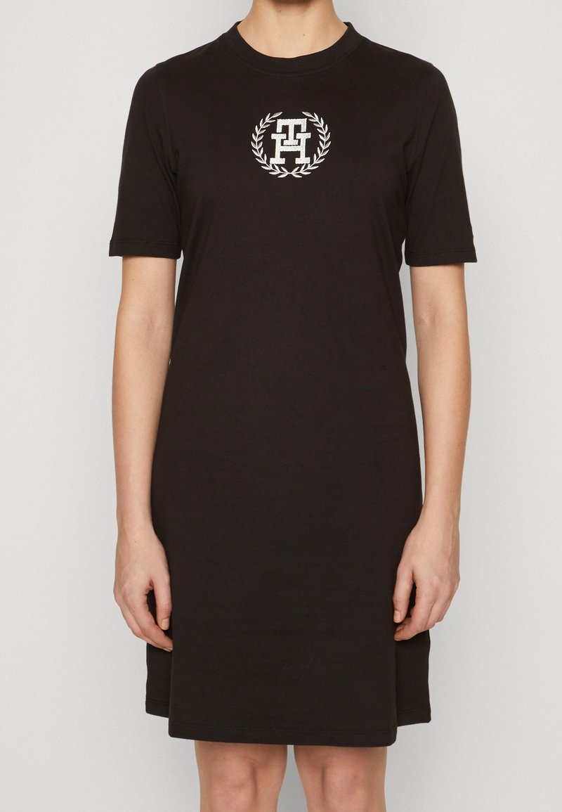 Robe noire en coton à manches courtes avec un col rond, présentant un design de logo blanc dans une couronne de laurier circulaire sur la poitrine.