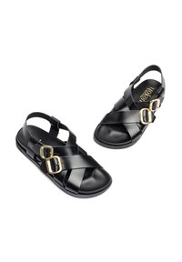 Schwarze Leder-Sandalen mit gekreuzten Riemen, zwei goldfarbigen Schnallen und einem strukturierten Fußbett. Offenes Design im Rücken für ein einfaches Anziehen.