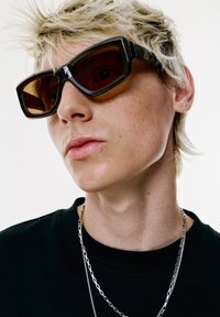 Mustat kulmikkaat aurinkolasit ruskeilla sävyillä, joita käyttää henkilö, jolla on vaaleat ja sotkuiset hiukset. Heillä on päällään musta paita ja hopeaketjuja.