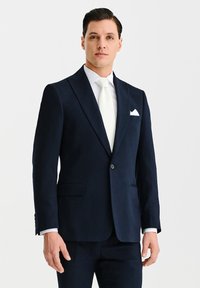 Granatowy blezer o smukłym kroju, z pojedynczym zapięciem, klapami typu notch oraz dwiema przednimi kieszeniami. Noszony z białym krawatem i poszetką.