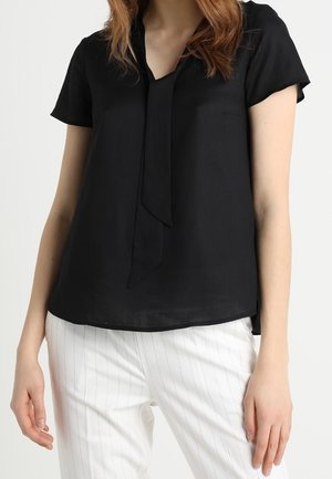 Vrouw draagt een zwarte blouse met korte mouwen en een strikdetail aan de voorkant, gecombineerd met witte broek met fijne strepen, tegen een effen achtergrond.