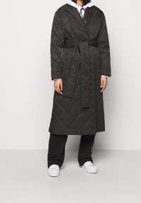 Osoba v dlouhém černém prošívaném kabátu s páskem, černých kalhotách, bílých teniskách a bílé mikině s černými šňůrkami.