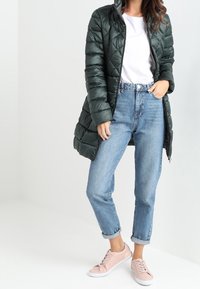 Abrigo acolchado largo y verde, camiseta blanca, vaqueros de cintura alta de color azul claro y zapatillas rosas. El abrigo cuenta con una cremallera y capucha.