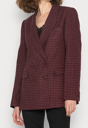 Vrouw draagt een dubbelgeknoopt bordeauxrood en zwart geruit blazer met klepzakken en een zwarte broek.
