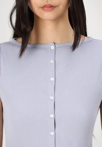 Lys lavendel ribbestrikket topp med rund hals og syv små hvite knapper foran, med korte ermer og en tettsittende stil.