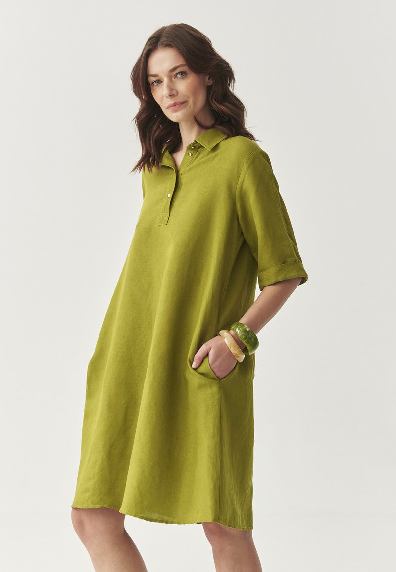 Rochie din in verde, cu guler, mâneci scurte și buzunare laterale. Prezintă accesorii contrastante, inclusiv brățări multicolore.