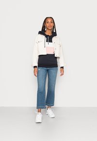 Jaqueta de ganga branca cortada, hoodie azul-marinho com riscas rosas e brancas, jeans azuis de boca de sino e ténis brancos. Design simples e ajuste casual.