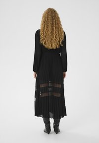 Vestido negro plisado con mangas largas, paneles transparentes en la parte inferior y cintura entallada, combinado con botas negras hasta la rodilla.