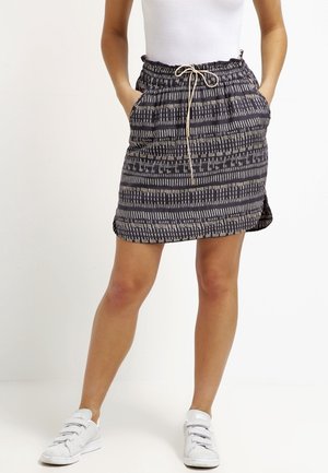 Femme portant une jupe à motifs noirs et blancs avec taille à cordon, débardeur blanc et baskets blanches, debout les mains dans les poches.