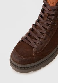Zapatilla alta de ante marrón con superficie texturizada, puntera redonda y cordones a juego. La suela de goma presenta un diseño estriado para mayor tracción.