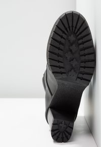 Bota negra con una suela de goma texturizada y un dibujo en el tacón, un tacón robusto y un upper liso. Diseño minimalista sobre un fondo claro.