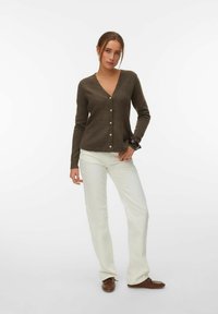 Frau trägt eine braune, geknöpfte Strickjacke, weiße Hose und braune Schuhe, steht mit einer Hand in der Tasche vor einem einfarbigen weißen Hintergrund.