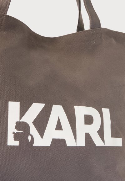Кафява текстилна торба с бели удебелени букви, изписващи KARL, включително силует на човек с конска опашка и слънчеви очила в буквата K.