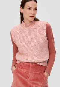Gestrickter ärmelloser Pullover in Hellrosa, mit hohem Kragen und strukturiertem Design, getragen über einem langärmeligen, rostfarbenen Oberteil und einer Hose aus geripptem Cord.