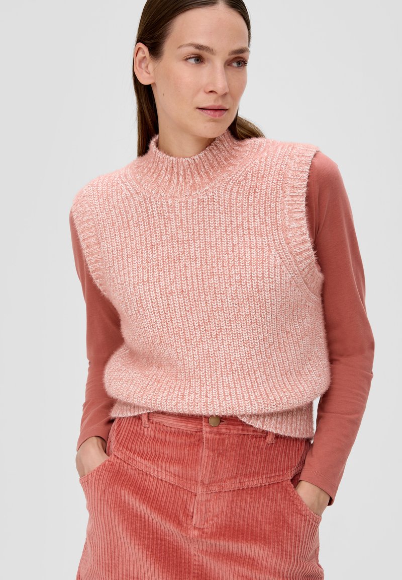 Gestrickter ärmelloser Pullover in Hellrosa, mit hohem Kragen und strukturiertem Design, getragen über einem langärmeligen, rostfarbenen Oberteil und einer Hose aus geripptem Cord.