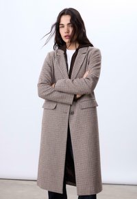Langmantel mit Hahnentwist-Muster in Braun, Beige und Creme. Ausgestattet mit einem Reverskragen, Vorderknöpfen und Seitentaschen. Mit Prinzessnähten.