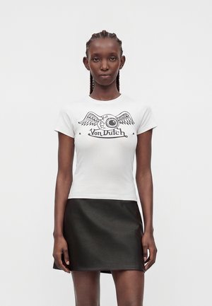 Jeune femme aux cheveux tressés portant un t-shirt blanc Von Dutch avec un motif d’œil ailé et une mini-jupe noire, debout devant un fond uni.