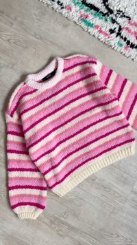 Πλεκτό πουλόβερ με ριγέ μοτίβο σε ροζ, λευκές και σκουρόροζ οριζόντιες ρίγες, απλωμένο επίπεδα πάνω σε ξύλινο πάτωμα δίπλα σε χαλί με σχέδια.