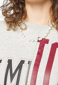 Collana di perle con forme e colori misti, esposta su una felpa grigia chiara con una stampa grafica rossa e nera.
