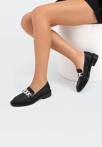 Sorte læder loafers med en sølv kæde accent, flad sål og glat tekstur. Designet med en rund tå og lav hæl.