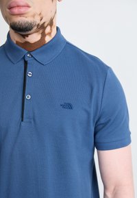 Modrá polo košile vyrobená z texturované látky s límcem, třemi knoflíky a malým vyšitým logem na levé straně hrudi.