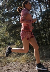 Atleta con giacca e pantaloncini rosa e bordeaux a motivo, che corre su un sentiero di terra. Indossa scarpe da corsa nere con suole bianche.