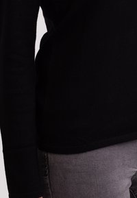 Svart stickad topp med långa ärmar, rund halsringning och croppad passform. Har en mjuk textur och diskreta sömdetaljer. Ihopparad med grå byxor.