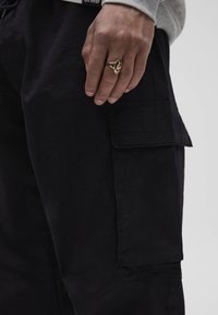 Czarne spodnie cargo mają boczną kieszeń, teksturowany materiał i luźny krój. Na spodniach spoczywa dłoń z złotym pierścionkiem.