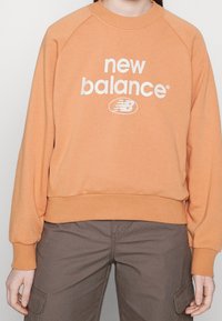 Felpa arancione con collo a girocollo a coste e maniche a sbuffo, con un grande logo bianco "new balance" sul davanti.