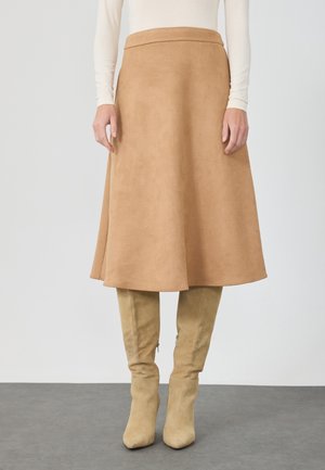 Kvinde iført beige A-formet midi-nederdel og matchende ruskindsstøvler til knæet, med en hvid langærmet top, står på en grå gulv.